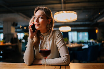 Portrait of beautiful blonde female in fashion clothes talking on smartphone with boyfriend, sitting in restaurant table with glass of red wine. Cute lady having phone conversation indoors in evening.