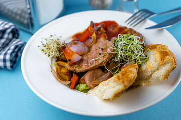 Warm salad with beef, vegetables, croutons and microgreen in a white plate on the restaurant table. Meat food in a bowl. Chop, medallions, cutlets. Blue background, close-up