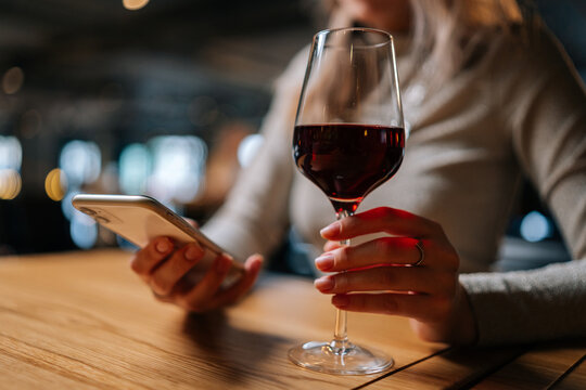 Close-up cropped shot of unrecognizable young woman using smartphone, typing online message sitting at table holding in hand glass of red wine at restaurant. Cute lady chatting with boyfriend. - Powered by Adobe