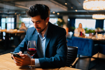 Portrait of handsome elegant man wearing fashion suit using smartphone chatting with friends and drinking red wine sitting at table in restaurant. Confident bearded male waiting for girlfriend on date