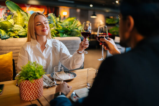 View From Back Of Unrecognizable Man In Suit Clinking Glasses Of Red Wine With Smiling Blonde Woman Sitting At Table In Fancy Restaurant At Evening. Happy Couple Enjoying Nice Romantic Dinner.