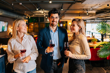 Portrait of cheerful man and two blonde women friends holding glasses of red wine, looking smiling at each other, standing posing in restaurant. Happy young male and female enjoying nice dinner.