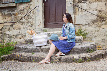 Attractive young woman sitting and posing with blue straw hat and big shopping bag on ancient stone stairs outdoor in old french city