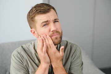 Dental health problems concept. Man suffering from strong toothache. Upset male patient with inflamed nerve, tooth abscess, or teeth sensitivity sitting on sofa and touching cheek with grimace of pain