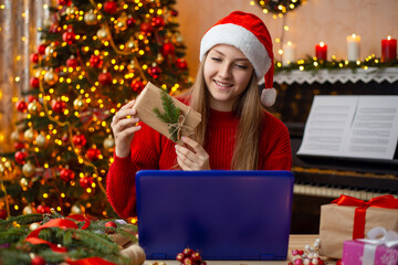 Young smiling blonde girl communicating with a friend via Internet, showing him a present. Happy teenage girl having online call with a friend who are far away, spending time together