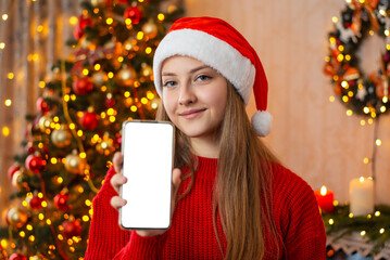 Girl showing screen of phone to the camera at home during Christmas celebration. Portrait of a young attractive girl with phone in decorated christmas room, wearing Santa hat