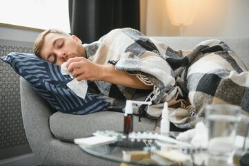 Sick bearded man who has bad cold or seasonal flu sitting on couch at home. Guy with fever wearing warm plaid shivering with worried face expression.