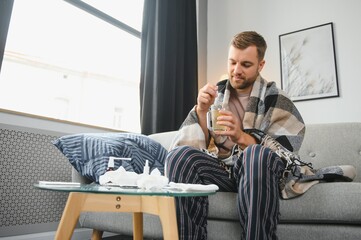 Sick bearded man who has bad cold or seasonal flu sitting on couch at home. Guy with fever wearing...