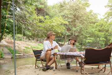 Pleased happy mother and daughter reading a book and using laptop while relaxing on the deck chairs in the river, sit near a camp and tent, drink coffee in a pine forest. Camping, recreation, hiking.

