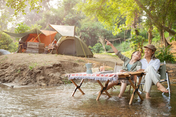 Pleased happy mother and daughter reading a book and using laptop while relaxing on the deck chairs in the river, sit near a camp and tent, drink coffee in a pine forest. Camping, recreation, hiking.