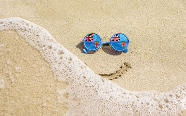 Sunglasses with flag of Fiji on a sandy beach. Nearby is a sea lightning and a painted smile. The concept of a successful vacation in the resorts of the Fiji.