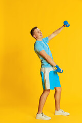 Fototapeta na wymiar Portrait of young man in blue uniform training, raising hands with dumbbells, posing isolated over yellow background. Concept of sport and emotions