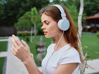 Woman wearing headphones listening to music through her phone smile with teeth walking in front of green palm trees on a summer trip
