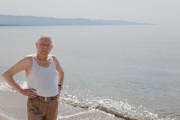Un signore anziano vestito in canottiera  in riva al mare si gode il sole estivo  - obrazy, fototapety, plakaty