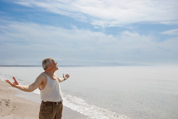 Un signore anziano vestito in canottiera  in riva al mare solleva le braccia al cielo 