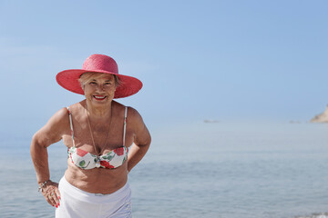signora anziana con il cappello colore fucsia si gode il sole  in riva al mare in costume da bagno - obrazy, fototapety, plakaty