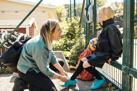 Side view of happy mother tying shoelace of son sitting on metal gate