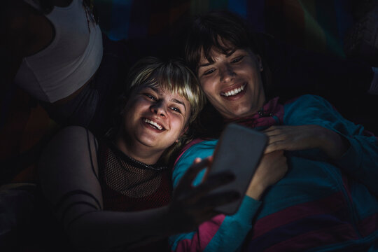 Happy non-binary person taking selfie with friend through mobile phone at night