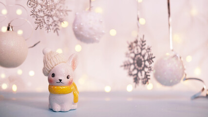 Christmas card, a small ceramic rabbit in a knitted hat and scarf stands against the background of Christmas balls and snowflakes. With blurred lights garlands.