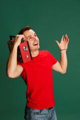 Portrait of young emotive man in casual clothes posing, listening to radio isolated over green background. Concept of emotions and music