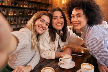 Three female friends take a selfie while having fun a drink in the cafeteria. Happy smiling Girls looking at the camera making grimace faces and having fun together. Millennial women and social media