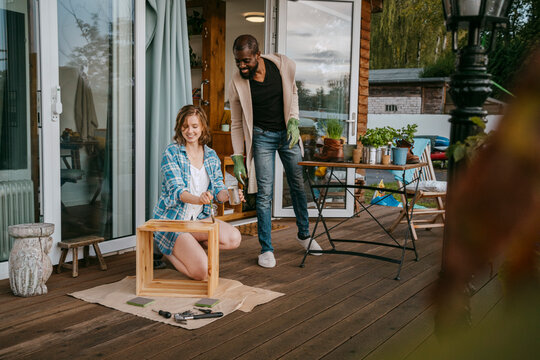 Mature man looking at woman painting furniture on porch outside house