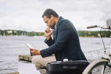 Side view of businessman looking at digital tablet while sitting on bench against bay