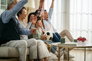 three generation asian family watching soccer game telecast on tv together at home