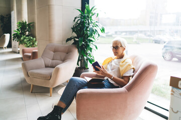 Woman sitting in armchair with book and smartphone in lobby