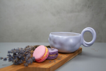 Light purple cup with coffee or milk, dessert macarons next to it, flowers, gray background