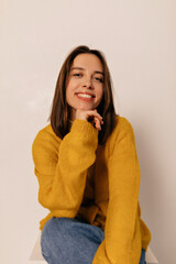 Happy young brunette woman in jeans and stylish yellow knitted shirt smiles sincerely, looks up and...