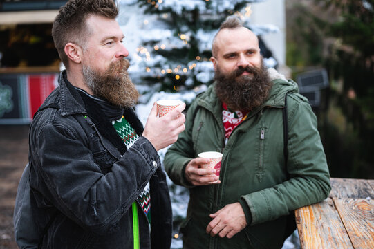 Long Beard Hipsters Homosexuals Enjoying Christmas Advent, Drinking Wine. Attractive Male Gay Couple Together For Xmas, Looking Away, Outdoor Shot