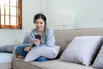 Asian woman listen music with mobile phone and headphones. Calm home weekend, relax while sit on sofa, enjoy favorite tune, popular music, use modern wireless headphones and gadgets in living room