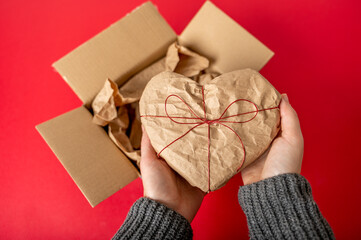Hands holding Heart-shaped gift box wrapped in kraft paper on red background. Just unpacked parcel
