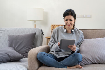 Happy pretty young woman in casual outfit sitting on sofa at home and using tablet while browsing internet