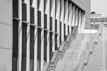 A wide empty staircase with stone and metal railings rises up among architectural panels and elements installed horizontally and vertically. Black and white photo