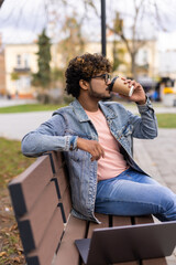 Indian young man sitting on a park bench and drinking coffee while working on laptop in outdoor fresh air at day time.