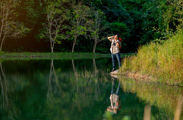 Fototapeta na wymiar Wide shot Asian man with bagpack and binoculars or other accessories for travel action of shout or bawl and stand near lake with reflection of his shadow during camping and travel in national park.