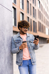 Young indian man standing in the street with a phone and coffee in his hands.