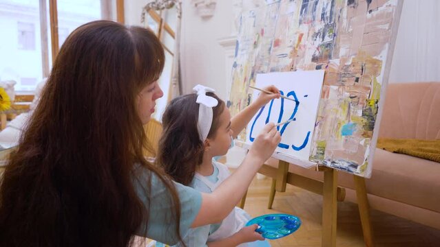 A girl with mom painting blue 2023 on an easel with canvas palette and brush.