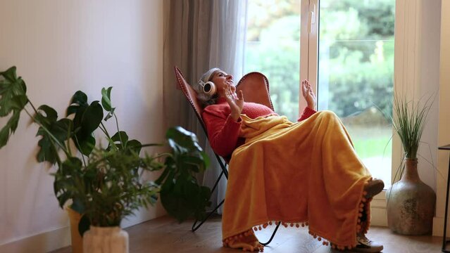 happy elderly woman sitting listening to music with headphones at home by the window, with plants.