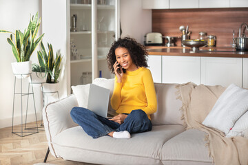 Distant work, working outside the office. Young African American woman freelancer or entrepreneur using laptop and mobile phone for work in home office sitting on sofa