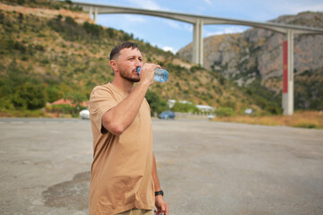 Obraz premium Confident young handsome man with beard drinking water from plastic bottle and enjoying mountains view at hot summer day. Healthcare concept. healthy lifestyle. Refreshing drink