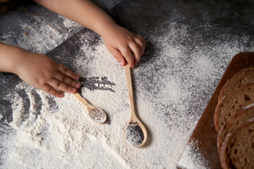 The hands of a kid who helps to make pastries . The child holds wooden spoons with seeds, flour is scattered on the table. High angle.