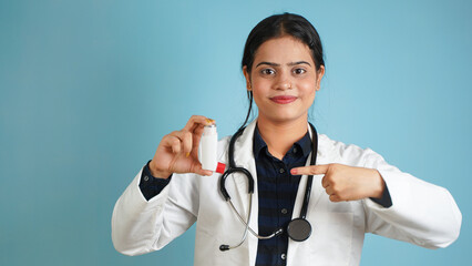 Portrait of a young female doctor wearing apron and stethoscope holding asthma inhaler, Cheerful Asian Indian woman doctor isolated over blue studio background - obrazy, fototapety, plakaty