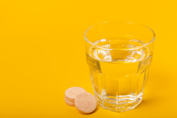 An effervescent vitamin tablet drops and dissolves in a glass of water on a yellow background. The concept of health. Medicine concept. Place for text.