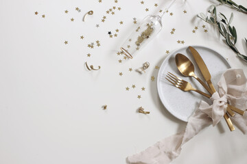 New Year festive still life. Birthday, wedding party. Celebration concept. Golden cutlery, beige silk ribbon on ceramic plate. Drinking glass, Golden star confetti isolated on white table background.
