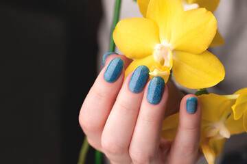 Female hand with beautiful manicure - blue glitter nails with yellow phalaenopsis orchid flower. Nail care concept