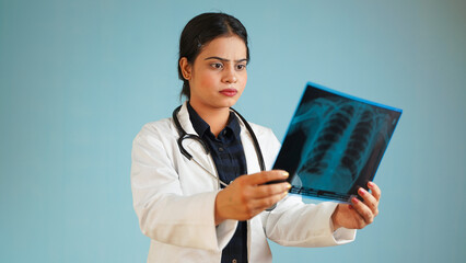 Portrait of a young female doctor examining patient's x-rays scan, Cheerful Asian Indian woman doctor in apron and stethoscope isolated over blue studio background