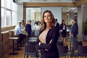 Fototapeta premium Portrait of confident and proud young woman against background of her office colleagues. Successful female company manager standing with arms crossed and looking at camera smiling. Confidence concept.